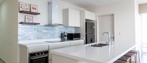 Una cocina moderna con armarios blancos, una isla grande con taburetes de bar, horno empotrado, microondas y un refrigerador de acero inoxidable.