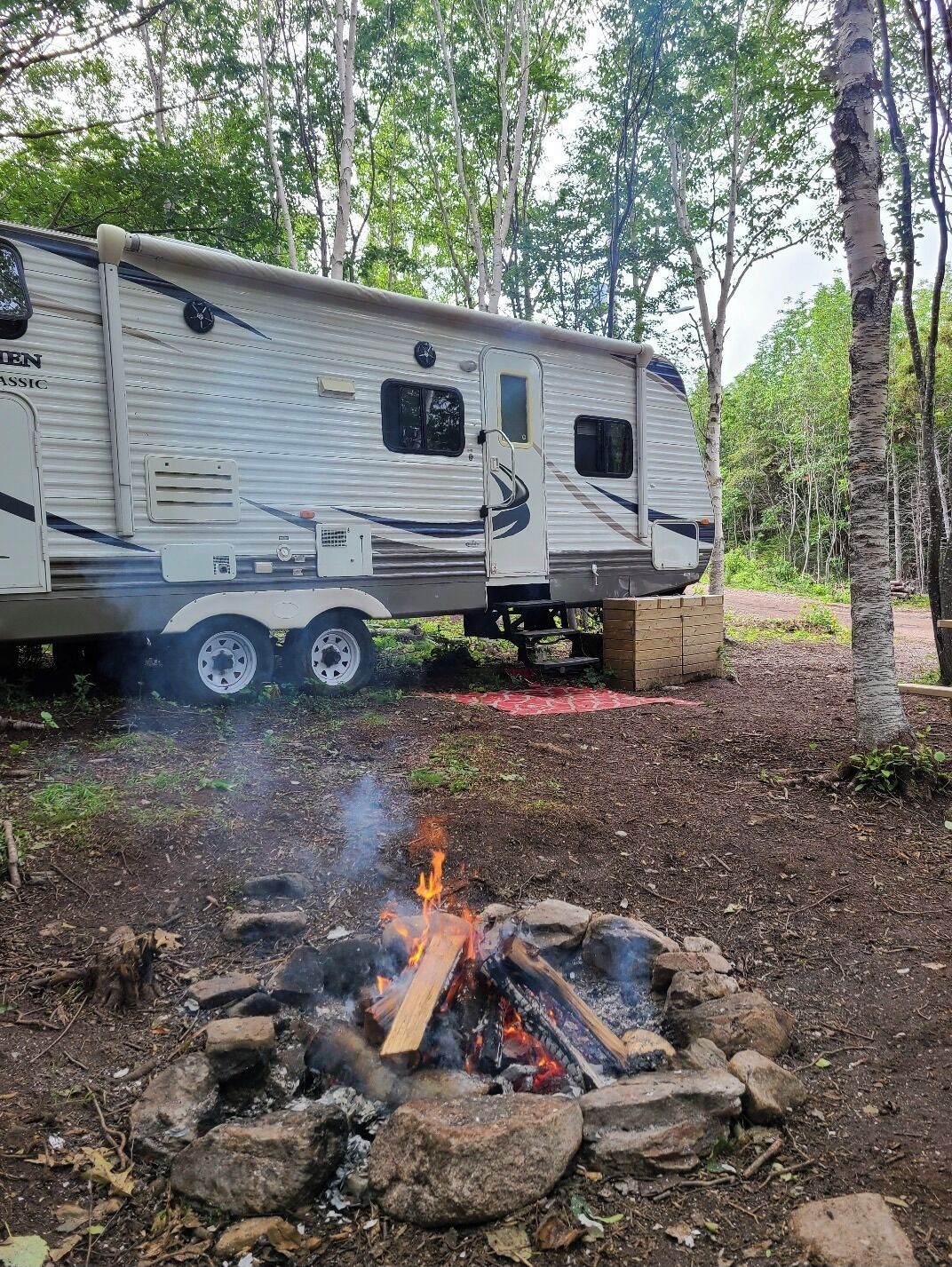 A recreational vehicle (RV) parked in a wooded area with a campfire in front.
