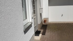 Una casa con fachada blanca, una ventana con marco blanco, una puerta blanca, un pequeño porche con reja de metal y una puerta de garaje blanca al fondo.