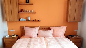 Un dormitorio con una cama de madera, dos mesitas de noche, una estantería con objetos decorativos y una pared grande de color naranja.