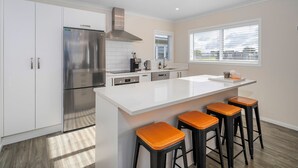A modern kitchen with white cabinetry, a stainless steel refrigerator, a built-in oven, and a large island with bar stools. - Cooks Family Retreat - Cooks Beach Holiday Home (Whitianga)