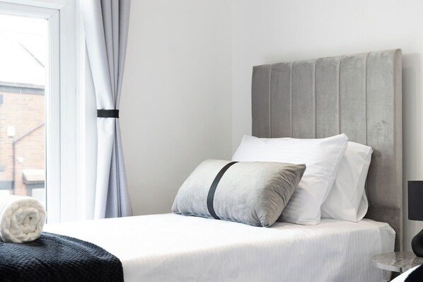 Una cama bien hecha con ropa de cama blanca y un cabecero gris, una toalla blanca enrollada a un lado y una ventana con cortinas.