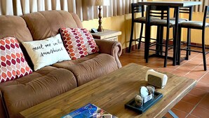 A living room with a brown sofa, patterned pillows, a wooden coffee table with magazines, a lamp, and a dining area with a table and chairs. - Marina Sol Resort Condo in Cabo
(Cabo San Lucas)