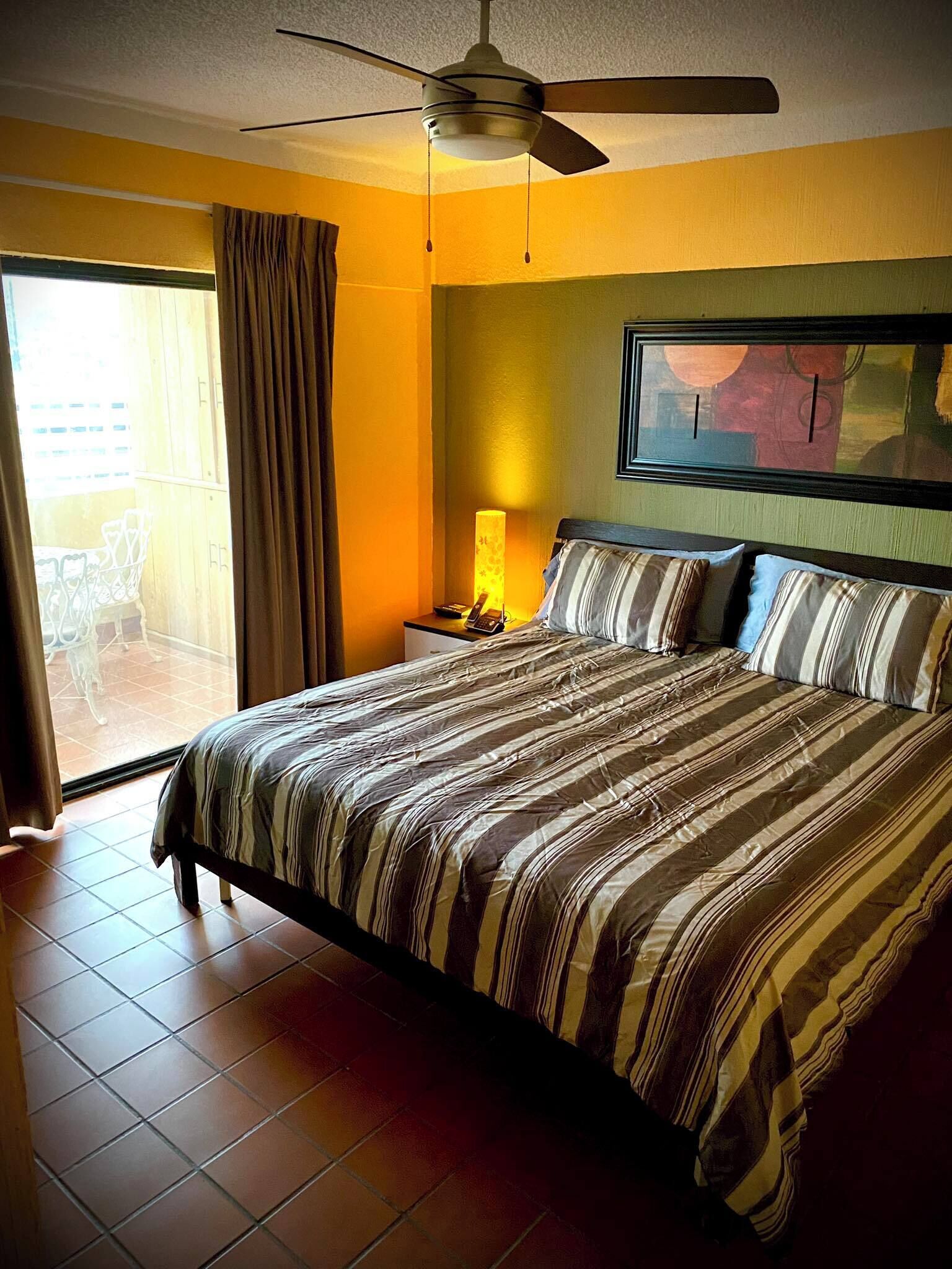 A hotel room with a bed, a ceiling fan, a nightstand, a lamp, and a view of the outside.