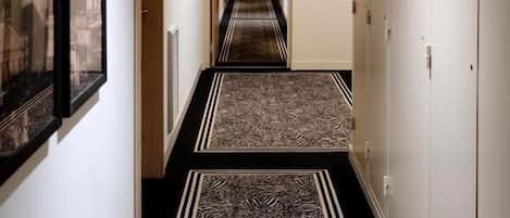 A hotel hallway with a zebra-striped carpet, framed pictures on the walls, and a row of lockers on one side.