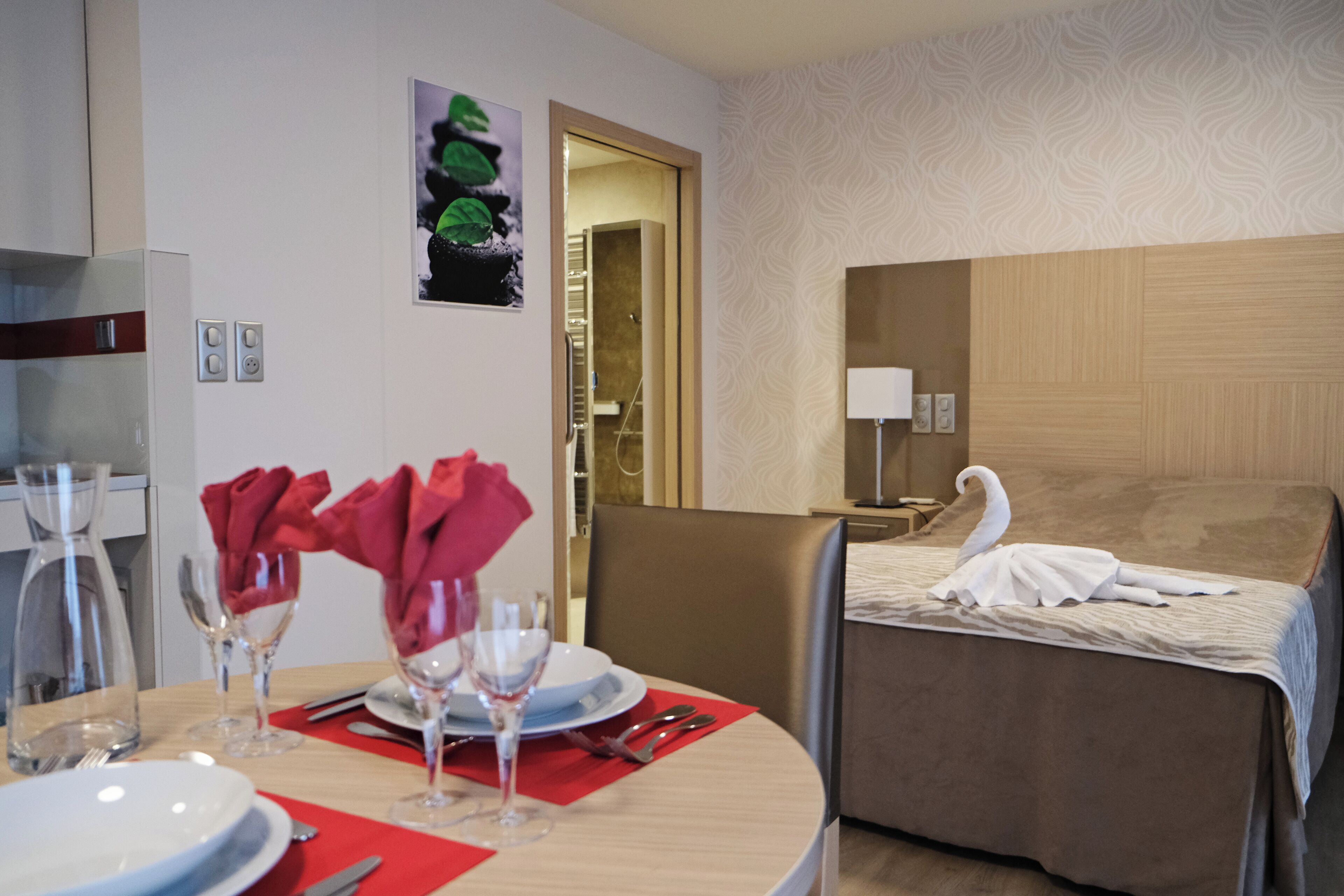 Ein Hotelzimmer mit Essbereich und Bett. Der Essbereich beinhaltet einen Tisch, der mit Tellern, Gläsern und roten Servietten gedeckt ist. Auf dem Bett liegen ein weißes Handtuch und eine Schwan-Dekoration.