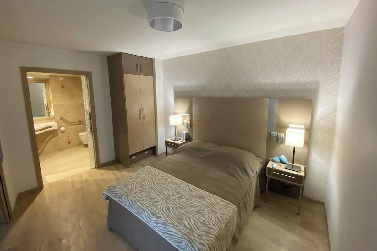 Une chambre d’hôtel avec un lit, une table de chevet, un bureau, une armoire et une salle de bain visible à travers une porte ouverte.