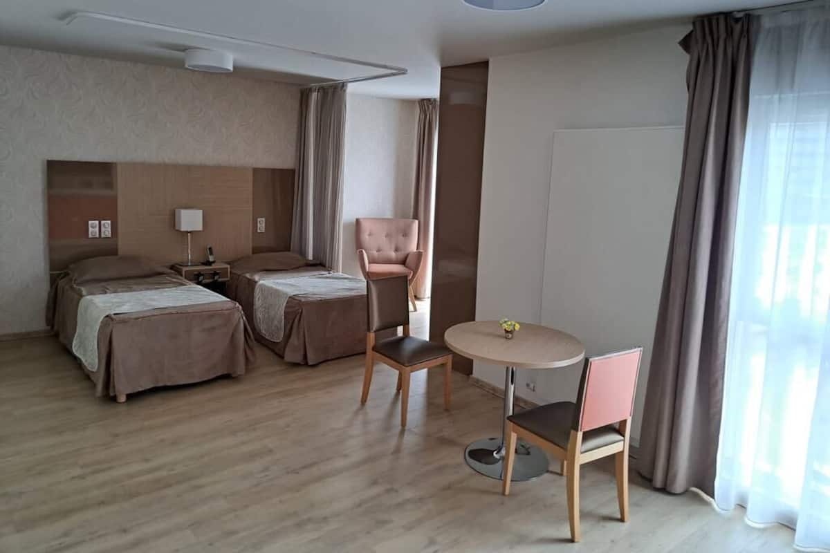 Une chambre d’hôtel avec deux lits, une petite table avec des chaises, une lampe et une vue sur l’extérieur grâce à de grandes fenêtres.