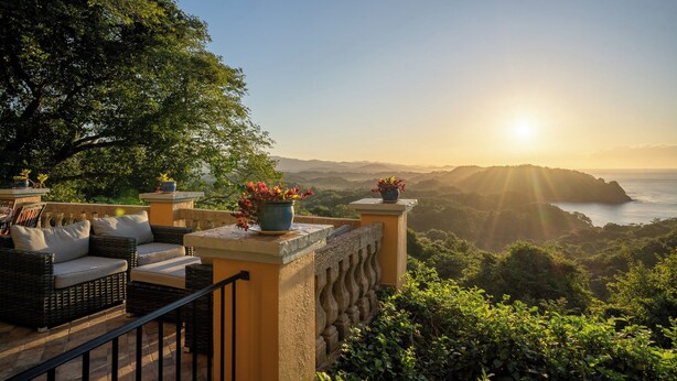 Une terrasse meublée de mobilier en osier et agrémentée de plantes en pot, offrant une vue sur un paysage montagneux et la mer au coucher du soleil.