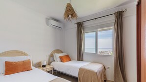 A hotel room with two beds, a window with a view of the sea, an air conditioning unit, and a hanging light fixture.