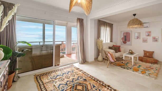 A spacious living room with a balcony, large windows, and a view of the sky.