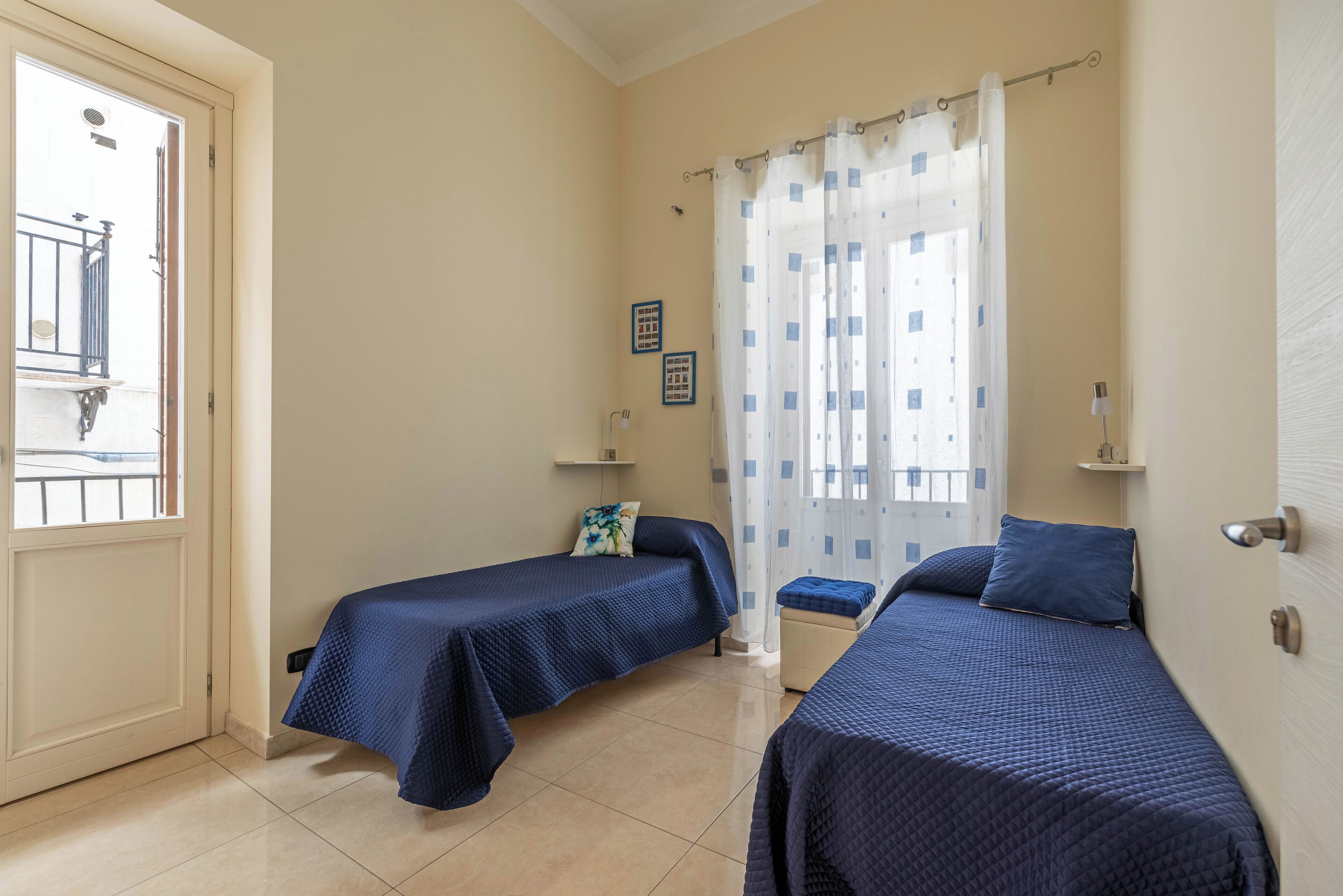 A room with two beds, a window with curtains, a door, and a small shelf with items.