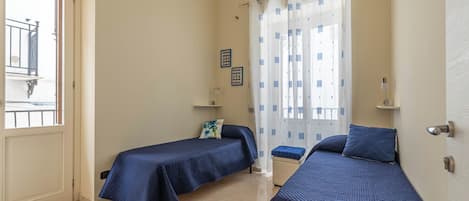 A room with two beds, a window with curtains, a door, and a small shelf with items.