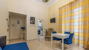A room with a bed, a dining table with chairs, a small table with a vase of flowers, a television on the wall, and a kitchen in the background. - Olivella Apartment al Teatro Massimo by Wonderful Italy (Palermo)