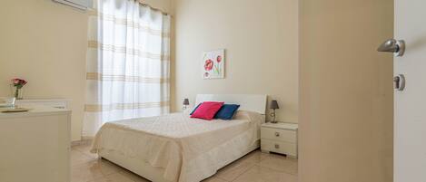 A bedroom with a bed, bedside tables, an air conditioning unit, a desk, and a window with curtains.