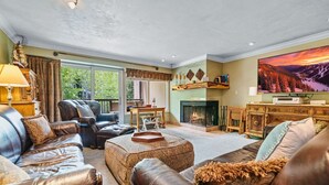 Ein geräumiges Wohnzimmer mit Ledersofas, einem Kamin, einem Fernseher und einem großen Fenster mit Blick auf Grünflächen.