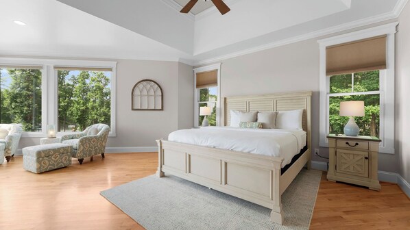 A spacious bedroom with a large bed, two armchairs, a nightstand, and a window with a view of trees.