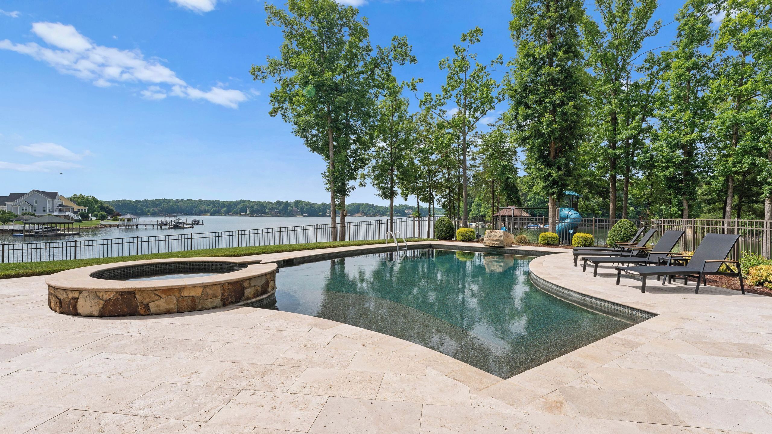 A swimming pool with a stone fire pit, lounge chairs, and a view of a lake.