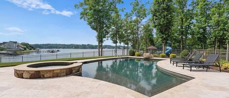A swimming pool with a stone fire pit, lounge chairs, and a view of a lake.
