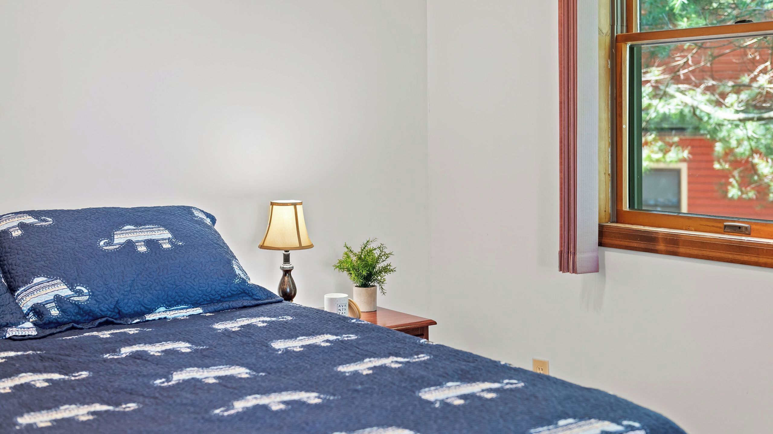 Un dormitorio con cama, mesita de noche con lámpara, una planta pequeña y ventana con vistas a la vegetación.