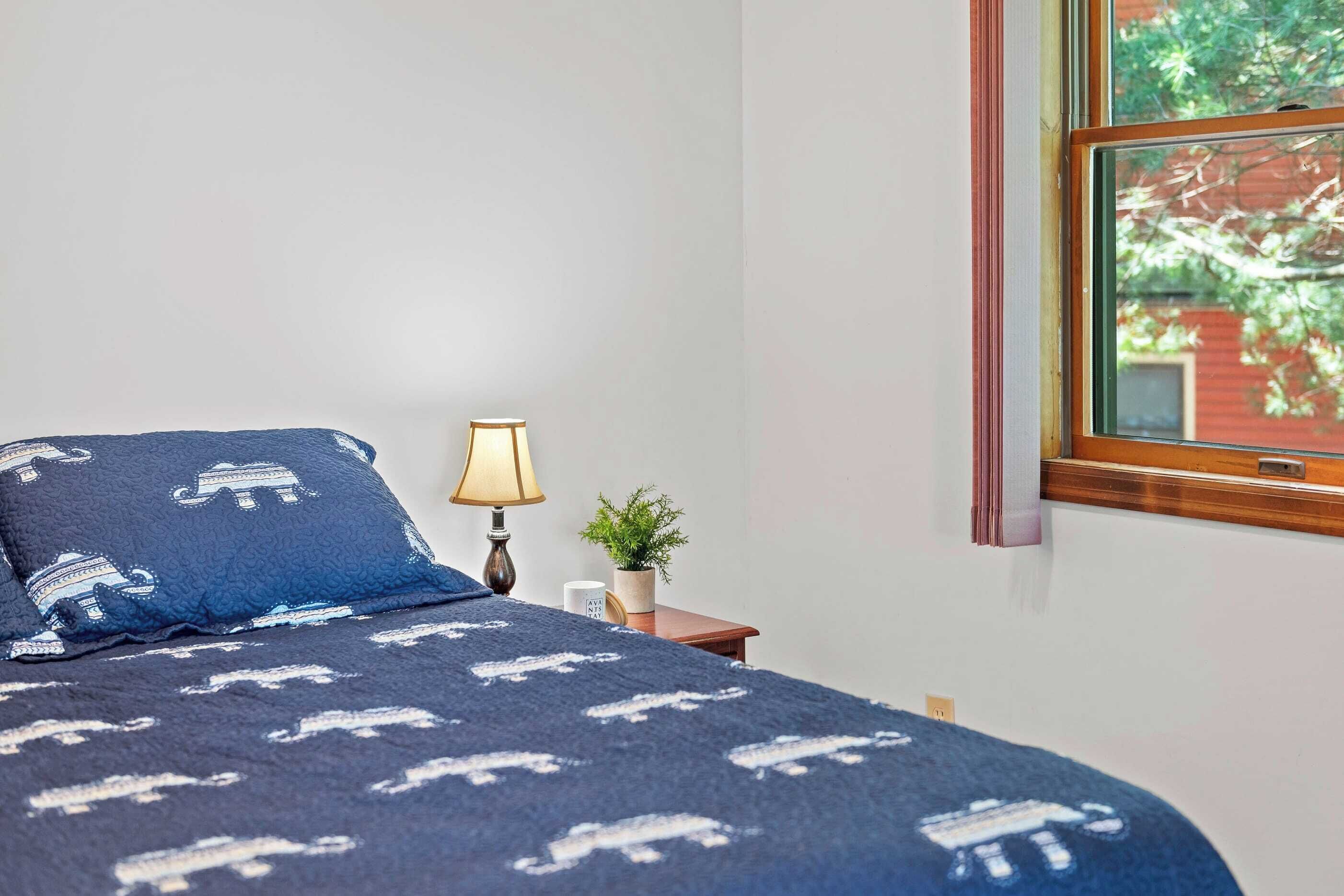 A bedroom with a bed, a lamp, a small plant, and a window with a view of trees.