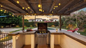 Area barbecue esterna con griglia, piscina e vista su case sullo sfondo.