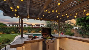 En udendørs terrasse med grill, pool og en træpergola med lyskæder.