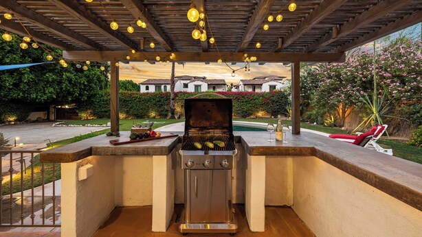 Et udendørs grillområde med grill, siddepladser og en pergola med hængende lys.