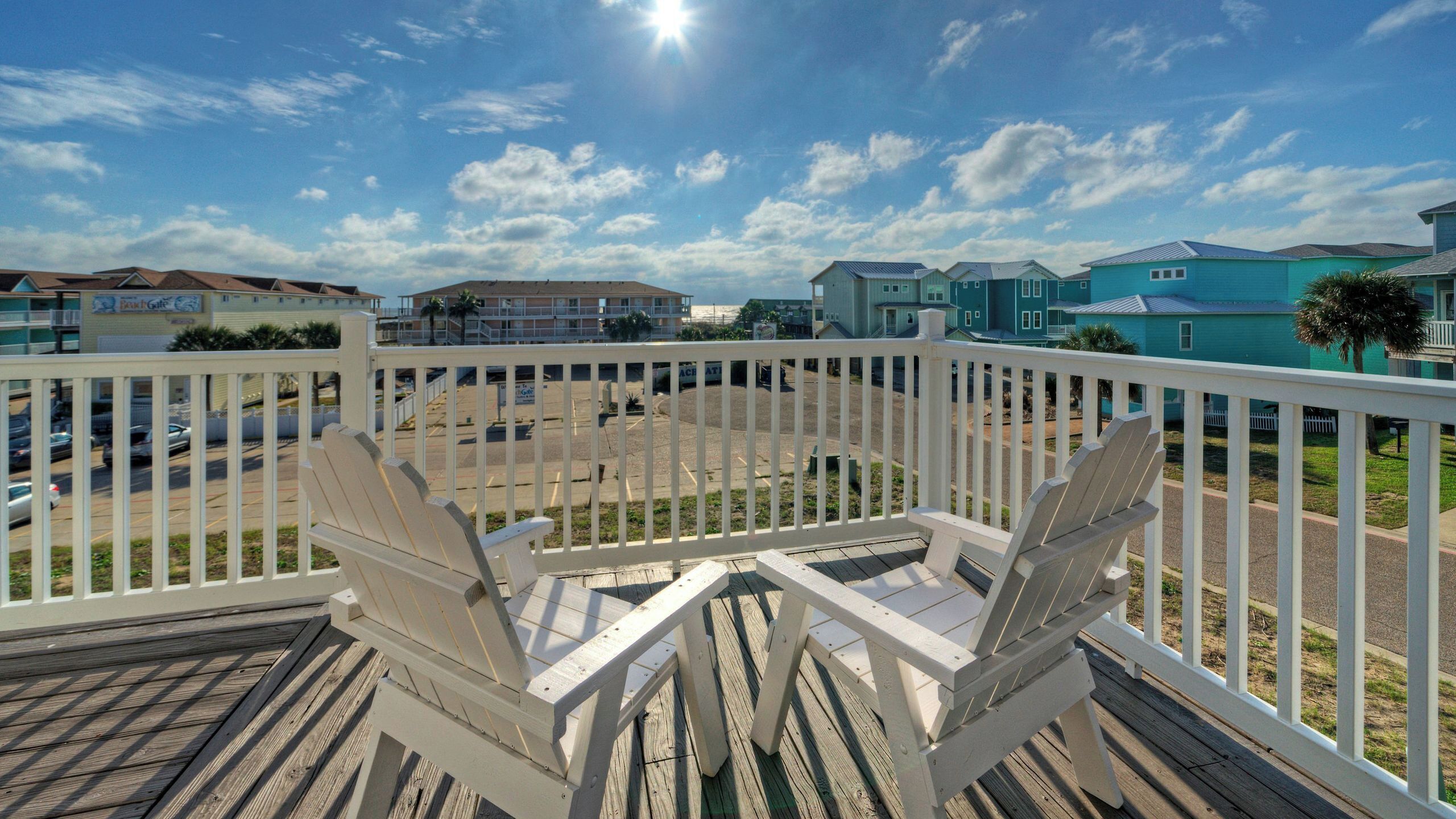 A wooden deck with two Adirondack chairs, a clear view of a parking lot, and buildings in the background.