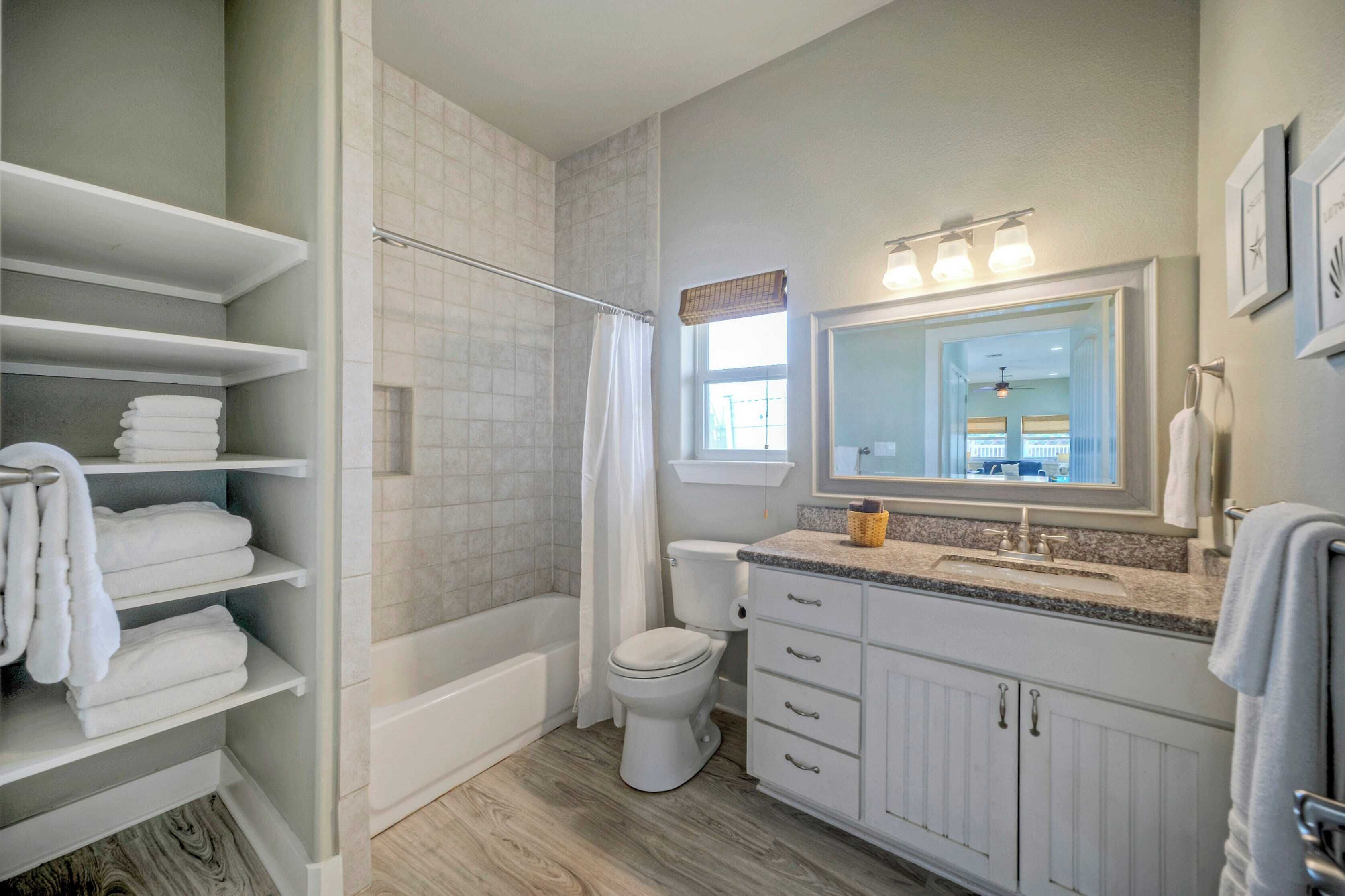 A bathroom with a bathtub, toilet, and vanity with a mirror. Shelves with towels are visible.