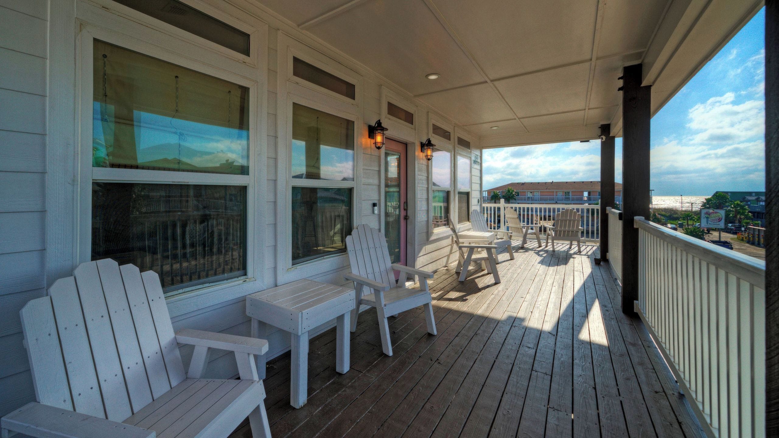 Eine überdachte Veranda mit weißen Adirondack-Stühlen, einem kleinen Tisch und Blick auf den Ozean.