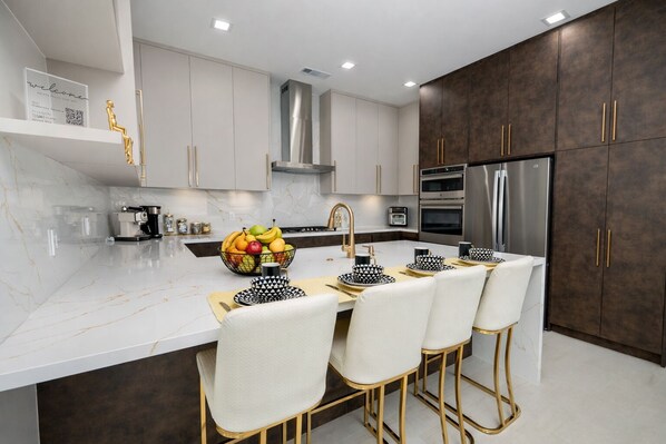 A modern kitchen with a marble countertop, stainless steel appliances, and a central island with bar stools. - The Hideaway Sleeps 23• Huge Glass Pool •Casio•Hike•Golf•Game Area (Indio)