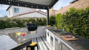 Cocina exterior con una parrilla, taburetes de bar y una zona de bar con bebidas.