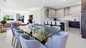 Una cocina moderna con gabinetes blancos, una isla central y un comedor con una mesa y sillas de vidrio.