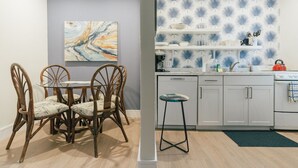 A dining area with a glass table and wicker chairs, a kitchen with white cabinets and blue patterned wallpaper, and a single stool.