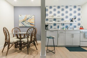A dining area with a glass table and wicker chairs, a kitchen with white cabinets and blue patterned wallpaper, and a single stool.