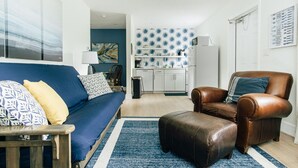 Ein modernes Wohnzimmer mit einem blauen Sofa, einem braunen Ledersessel und passendem Hocker. Im Hintergrund ist eine Küche mit weißen Schränken und einer blau gemusterten Spritzschutzwand zu sehen.