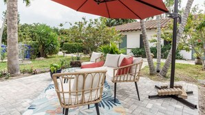 Eine Terrasse mit Korbmöbeln, einem roten Sonnenschirm und einem kleinen Garten mit Palmen und Topfpflanzen.