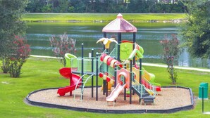 Un parque infantil con toboganes y estructuras para escalar, rodeado de césped y una masa de agua.