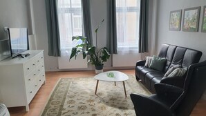 Ein Wohnzimmer mit einer Couch, einem Couchtisch, einem Fernseher auf einem Schrank, einer Topfpflanze und einem Fenster mit Vorhängen.