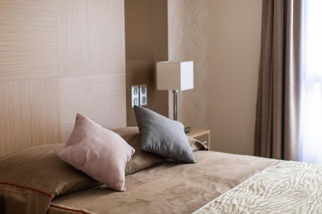 Habitación de hotel con cama, almohadas, mesita de noche, lámpara y ventana con cortinas.