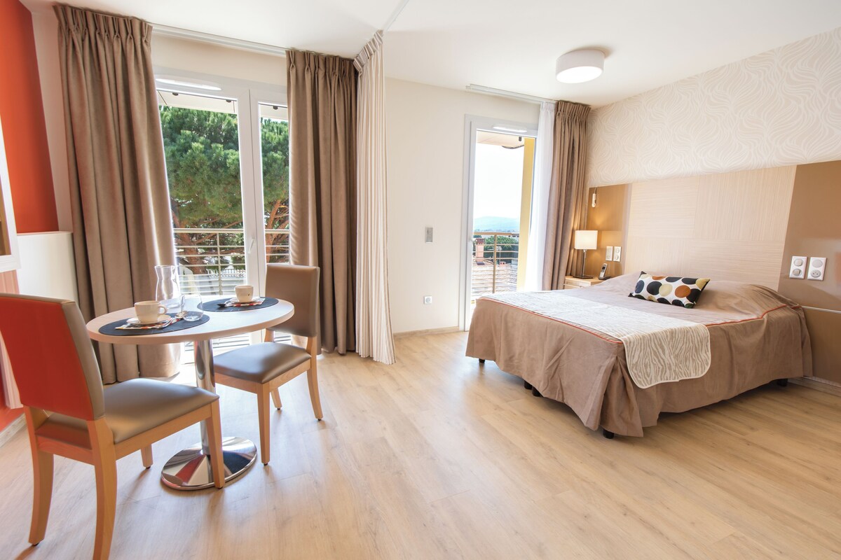 A hotel room with a bed, a dining table with chairs, a balcony with curtains, and a view of the outdoors.