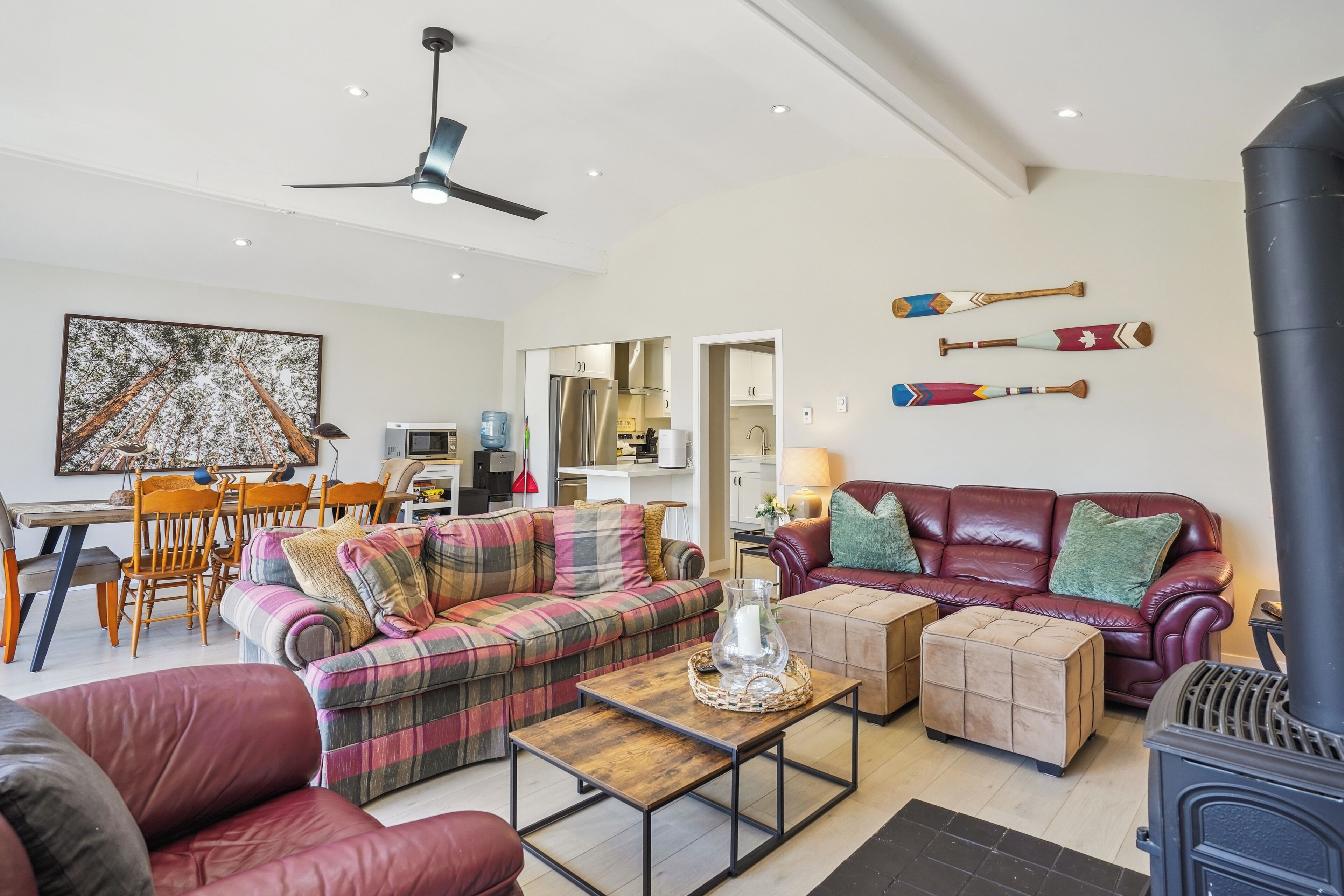 A living room with a plaid sofa, ottoman, and a coffee table. There is a fireplace, a ceiling fan, and a dining area in the background.
