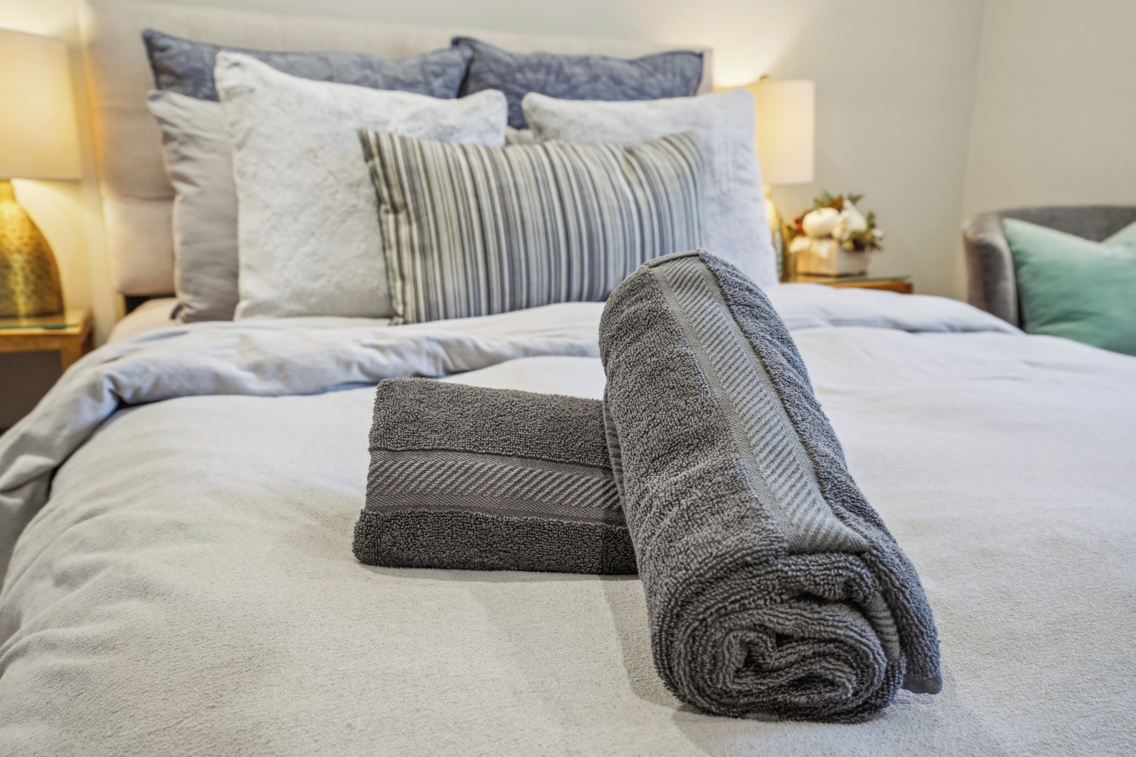 Una cama bien hecha con una toalla gris y un edredón doblado, una almohada a rayas y una lámpara en la mesita de noche.