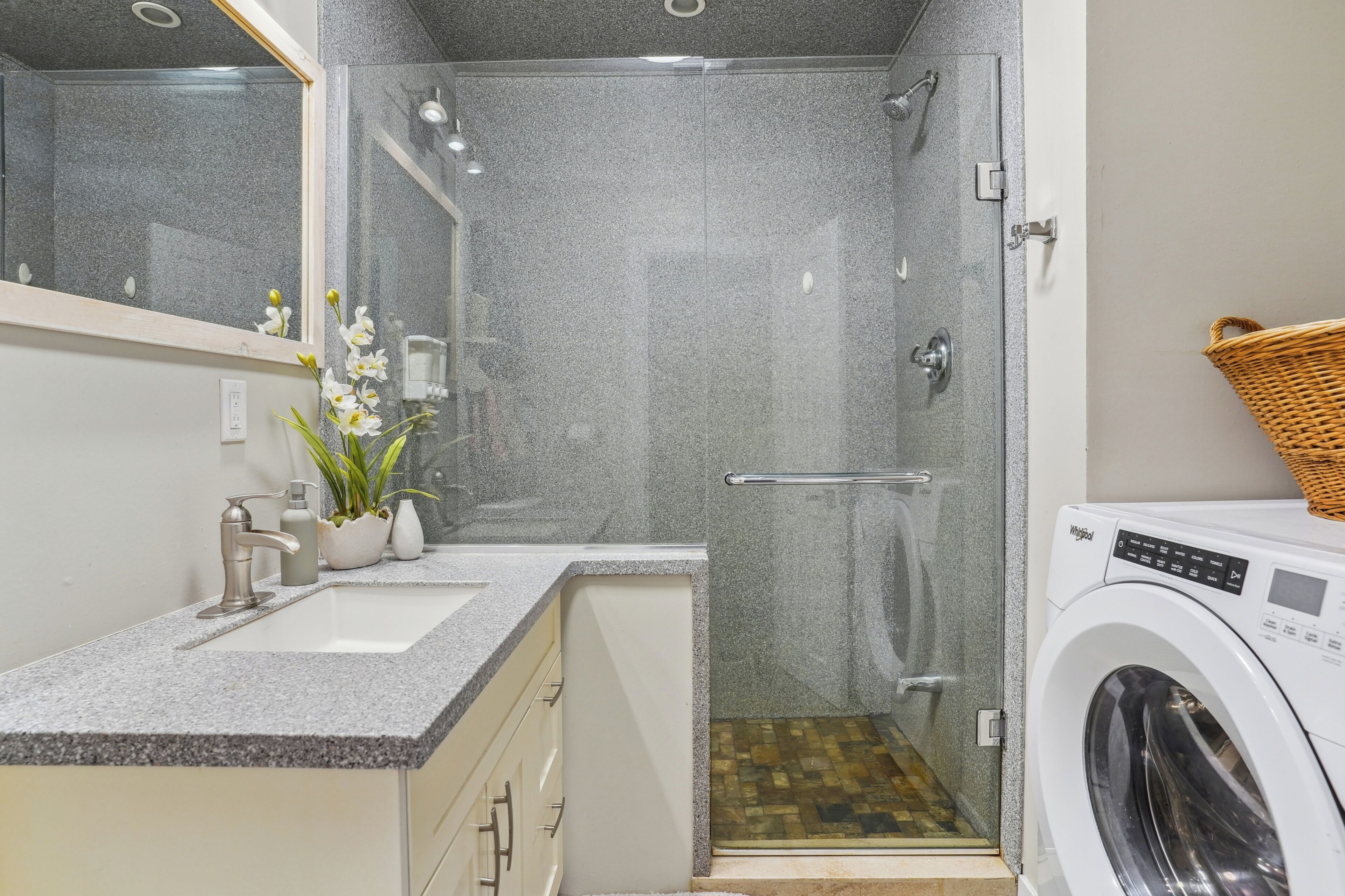 A modern bathroom with a granite countertop, a sink, a mirror, a shower enclosure, and a washing machine.