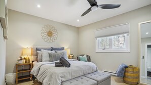 Une chambre à coucher comprenant un lit, des tables de chevet, un ventilateur de plafond, une fenêtre avec des stores et un panier.
