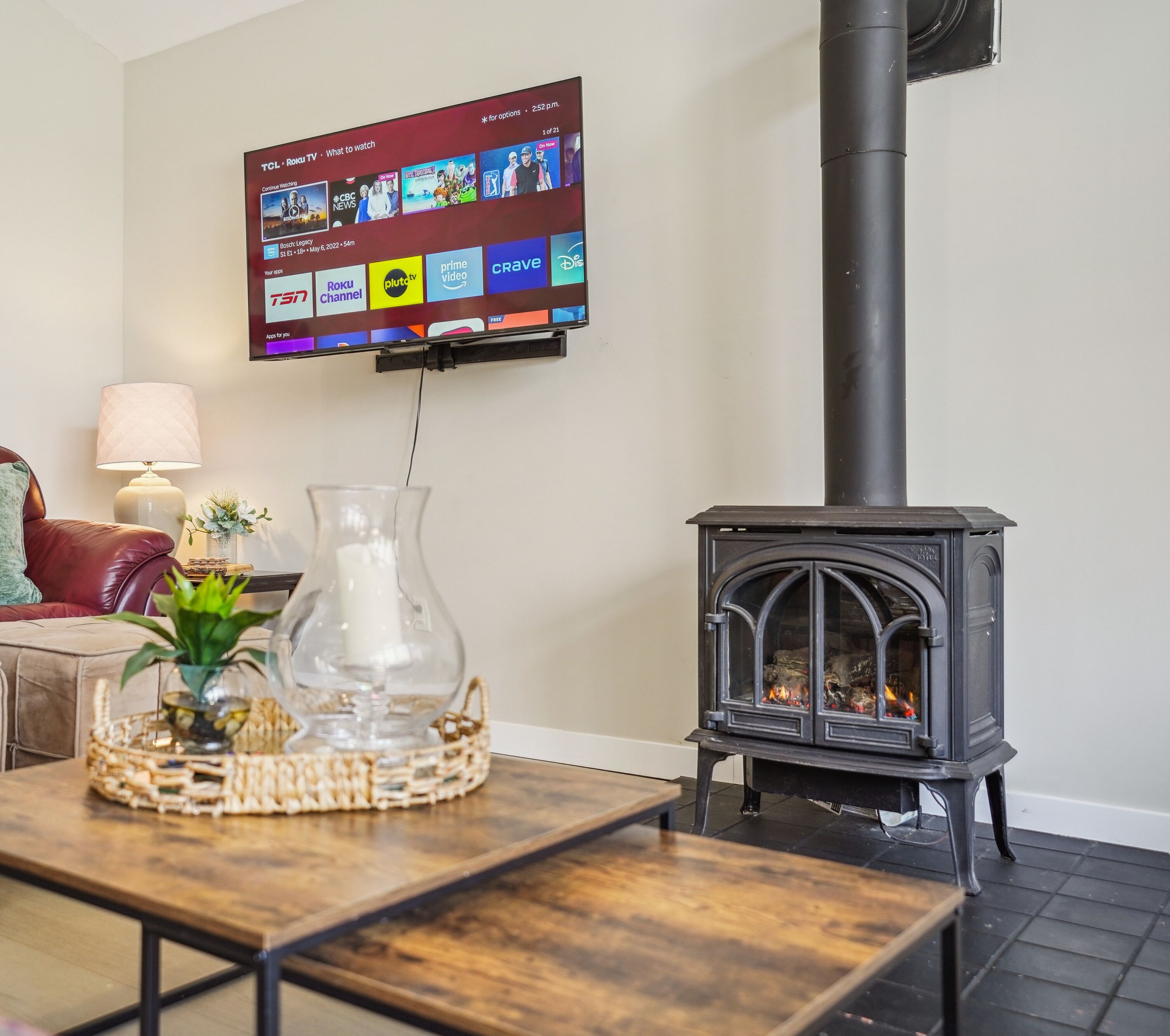 A living room with a television mounted on the wall displaying various channels, a black wood-burning stove, a wooden table with a vase and a plant, and a red leather sofa.