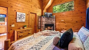 Une chambre confortable dans un chalet, avec une cheminée en pierre, des murs en bois, un lit recouvert d’une literie à motifs floraux et une fenêtre donnant sur la verdure.