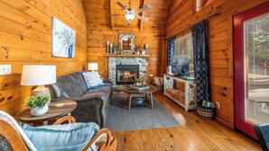 L’intérieur chaleureux d’un chalet avec une cheminée, des murs en bois, un ventilateur de plafond et un coin salon confortable.