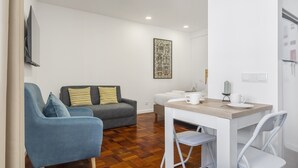 A compact living space with a sofa, a dining table with chairs, a flat-screen TV mounted on the wall, and a decorative wall piece. - Ribeira do Esmeraldo 3B by Madeira Sun Travel (Funchal)